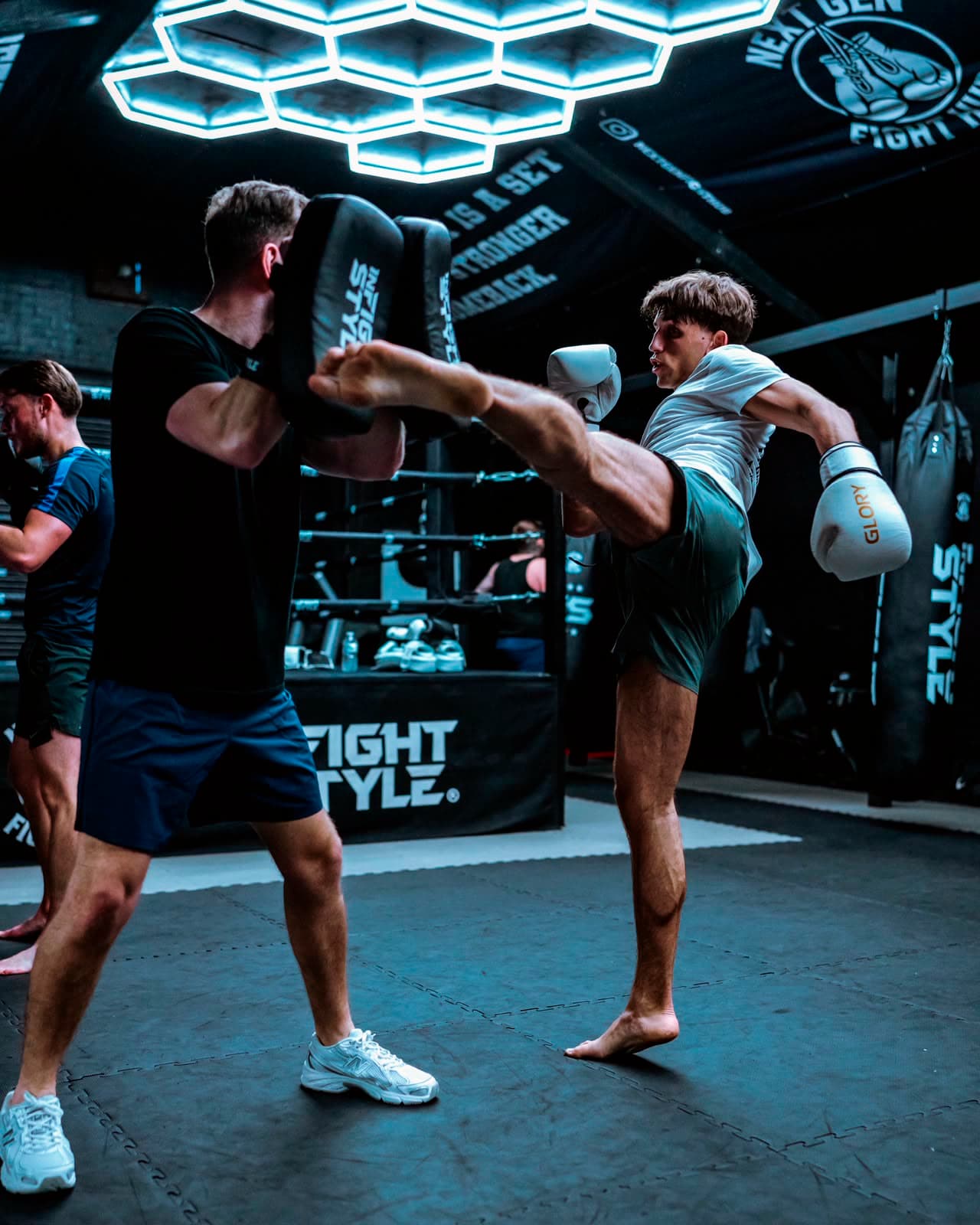 Head coach Hayden holding the kick shield for a fighter throwing a head kick under the hexagon lights